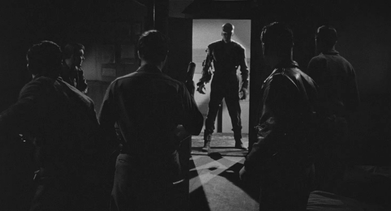 5 men in a darkened room confront The Thing monster as it emerges through a lit doorway